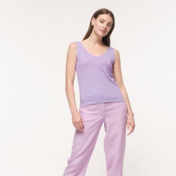 Frau in einem lila Top und rosa Hose steht vor einem weißen Hintergrund.