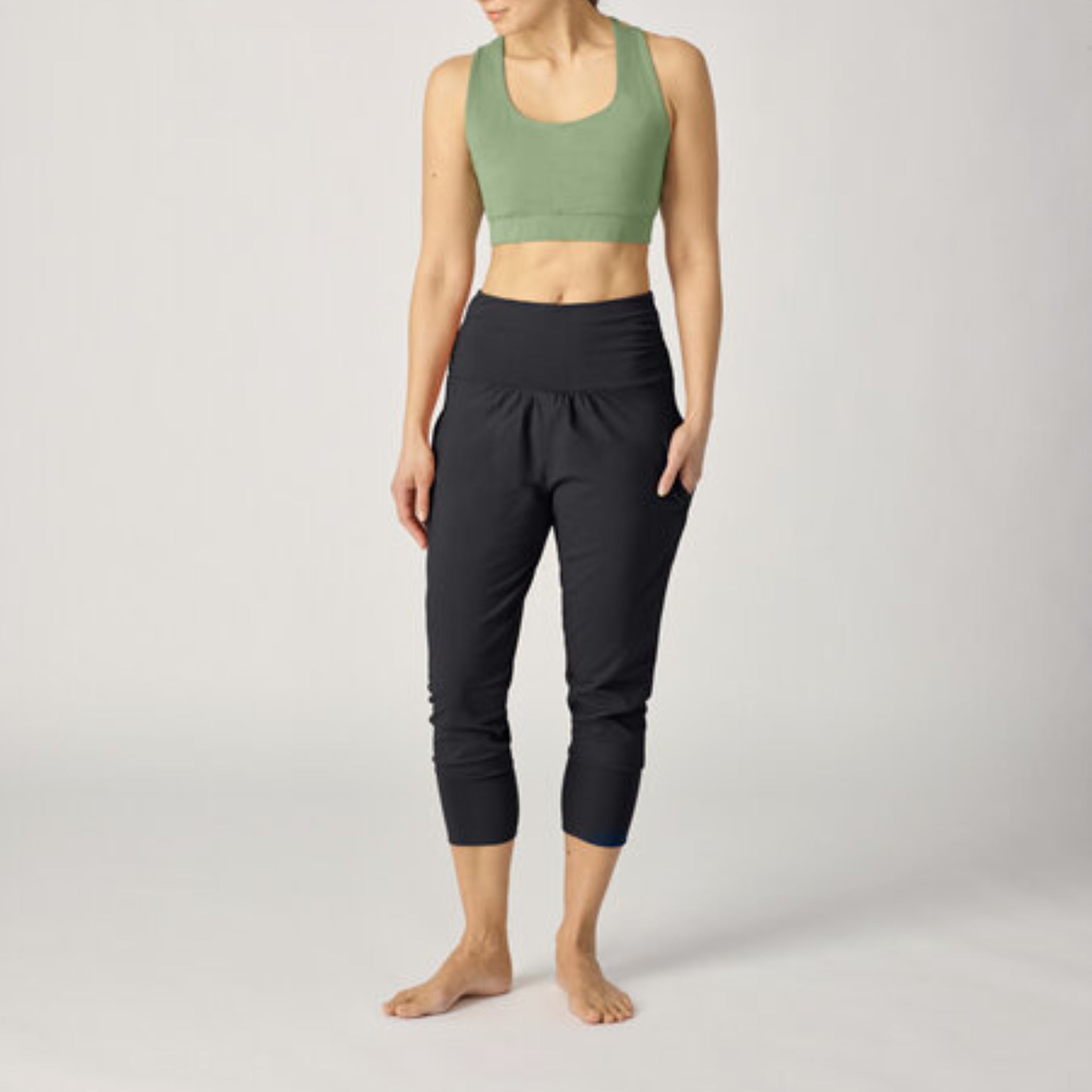 Frau in grünem Sport-BH und schwarzen Yoga-Hosen steht barfuß auf hellem Hintergrund.