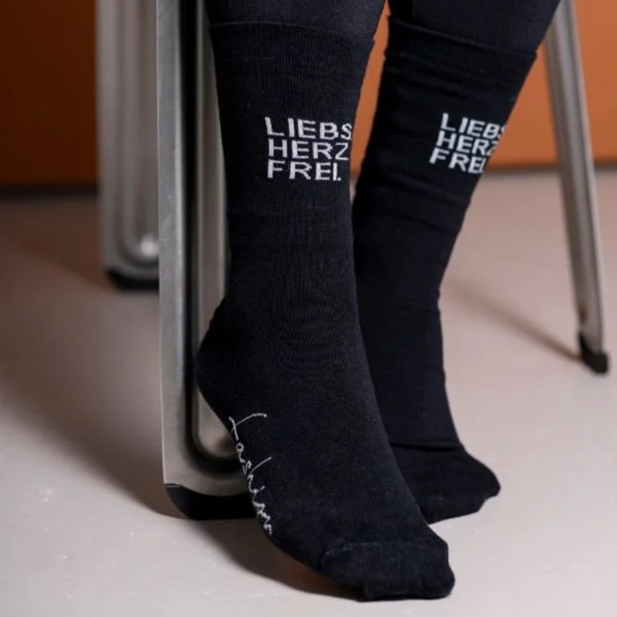 Eine Person, die schwarze Socken mit der weißen Aufschrift "LIEBS HERZ FREI" trägt, sitzt mit gekreuzten Füßen unter einem Stuhl auf einem hellen Boden. Ein weiteres Wort in Schreibschrift ist an der Seite einer Socke zu sehen.