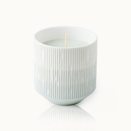 Weiße Kerze in einem strukturierten, zylindrischen Gefäß mit vertikalen Linien auf hellem Hintergrund.