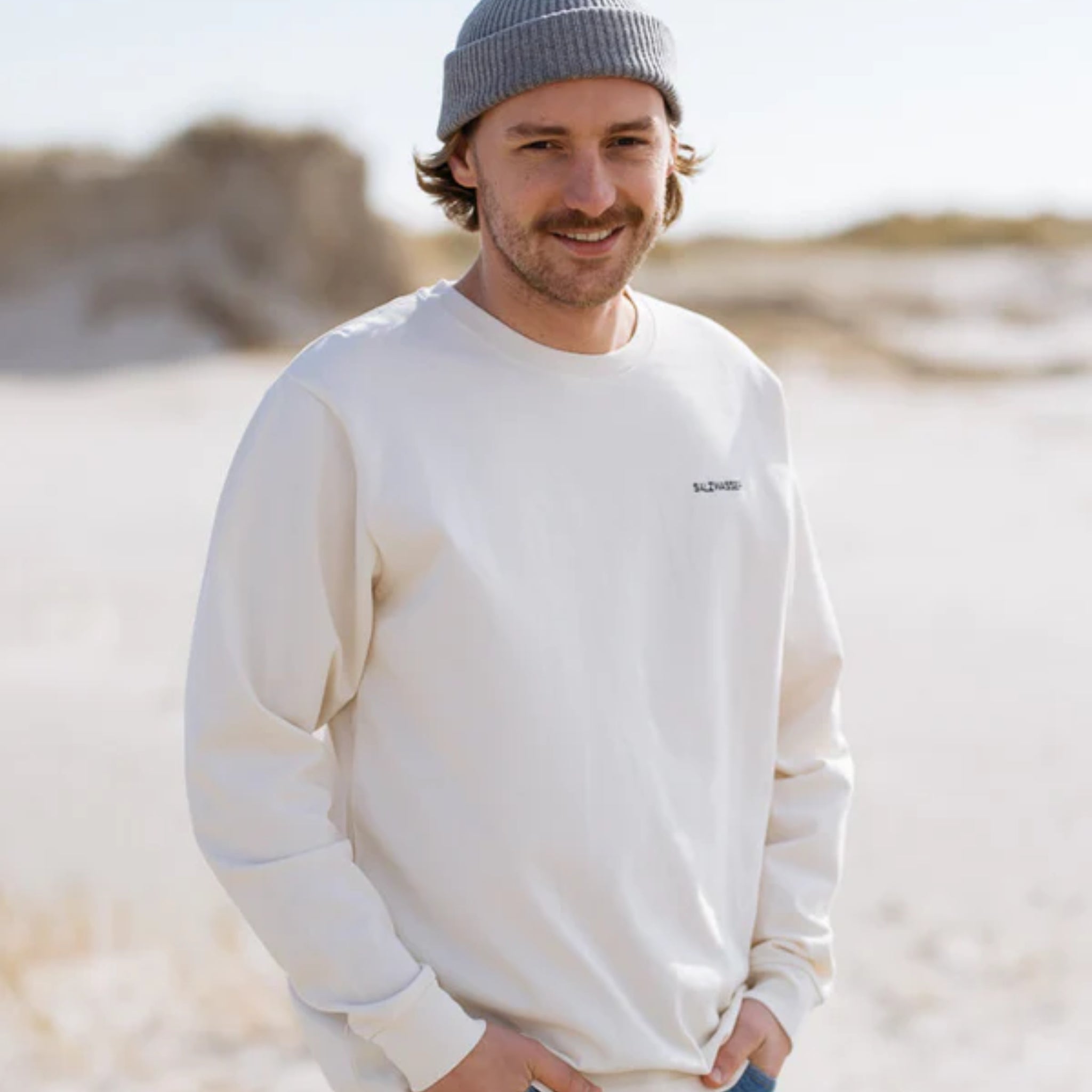 Person mit Mütze und weißem Sweatshirt steht lächelnd am Strand, Hintergrund unscharf und sandig.