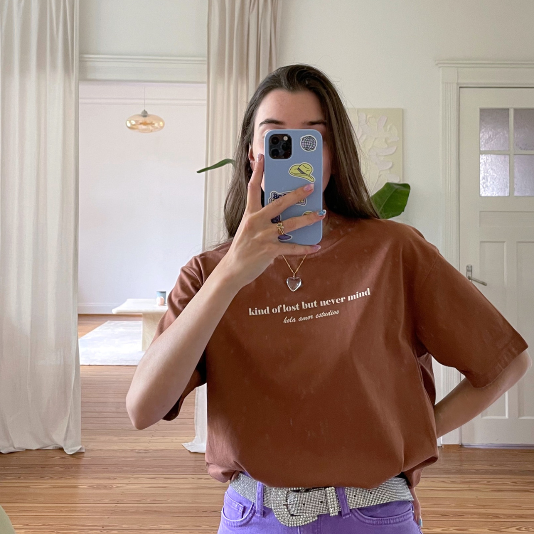 Eine Person mit langen braunen Haaren macht ein Spiegel-Selfie in einem hellen Raum. Sie trägt ein braunes T-Shirt mit weißem Text, eine lila Hose und einen glitzernden Gürtel. Ihre Handytasche hat Aufkleber, und im Hintergrund hängen helle Vorhänge.