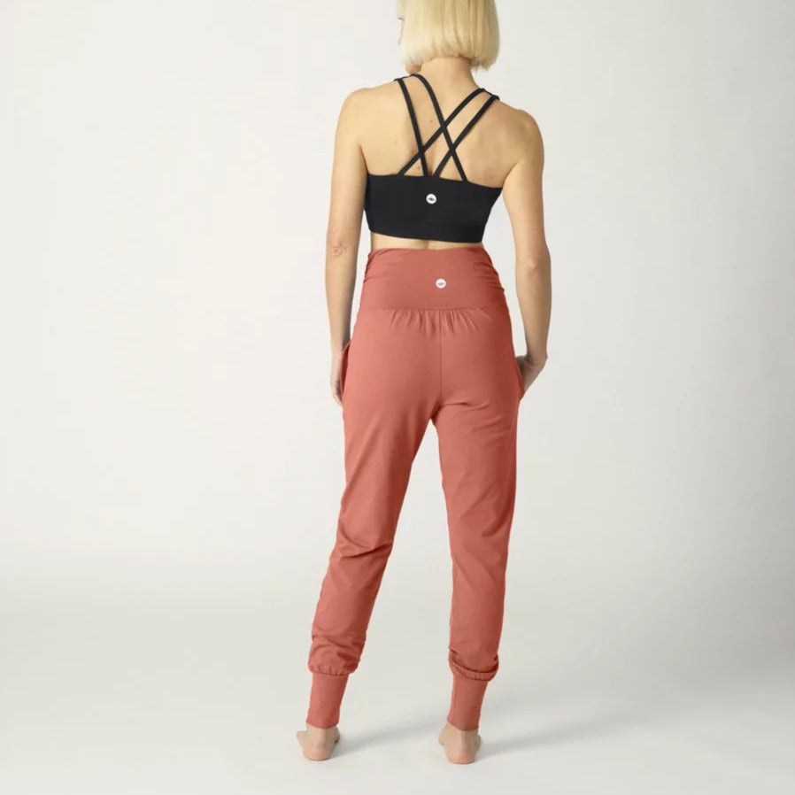 Eine Frau mit blonden Haaren trägt ein schwarzes Trägershirt und rostfarbene Hosen, stehend von hinten.