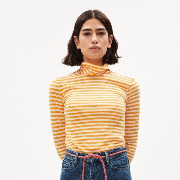 Frau mit schulterlangem Haar trägt ein gelb-weiß gestreiftes Rollkragenshirt und blaue Jeans vor weißem Hintergrund.