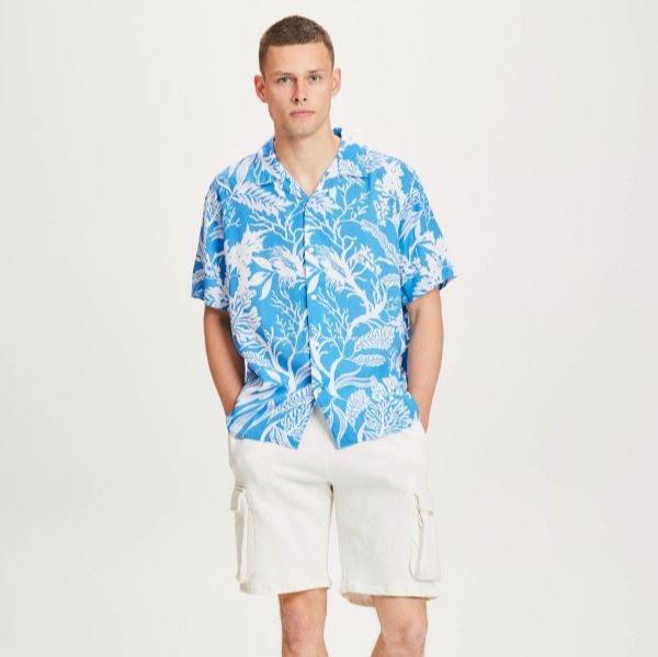 Ein Mann trägt ein blaues Hawaiihemd und weiße Shorts, steht vor einem weißen Hintergrund.