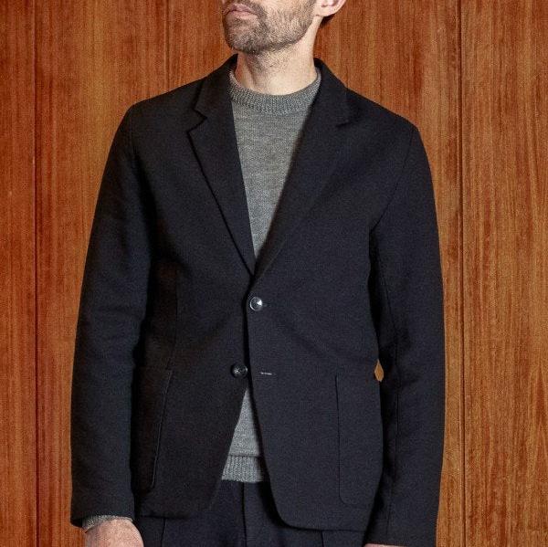 Ein Mann trägt ein schwarzes Sakko über einem grauen Pullover vor einem hölzernen Hintergrund.