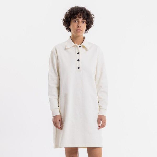 Person in a cream shirt-dress with black buttons stands against a plain white background.