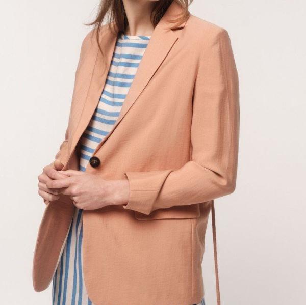 Frau trägt einen apricotfarbenen Blazer über einem blau-weiß gestreiften Kleid, vor weißem Hintergrund.