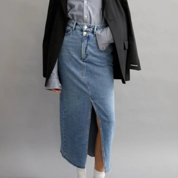 Eine Frau steht vor einem einfarbig grauen Hintergrund. Sie trägt einen schwarzen Blazer über einem gestreiften Hemd und einen langen Jeansrock mit einem vorderen Schlitz, gepaart mit weißen Socken und schwarzen Slippern. Ihr Haar ist offen und sie hat einen neutralen Gesichtsausdruck.