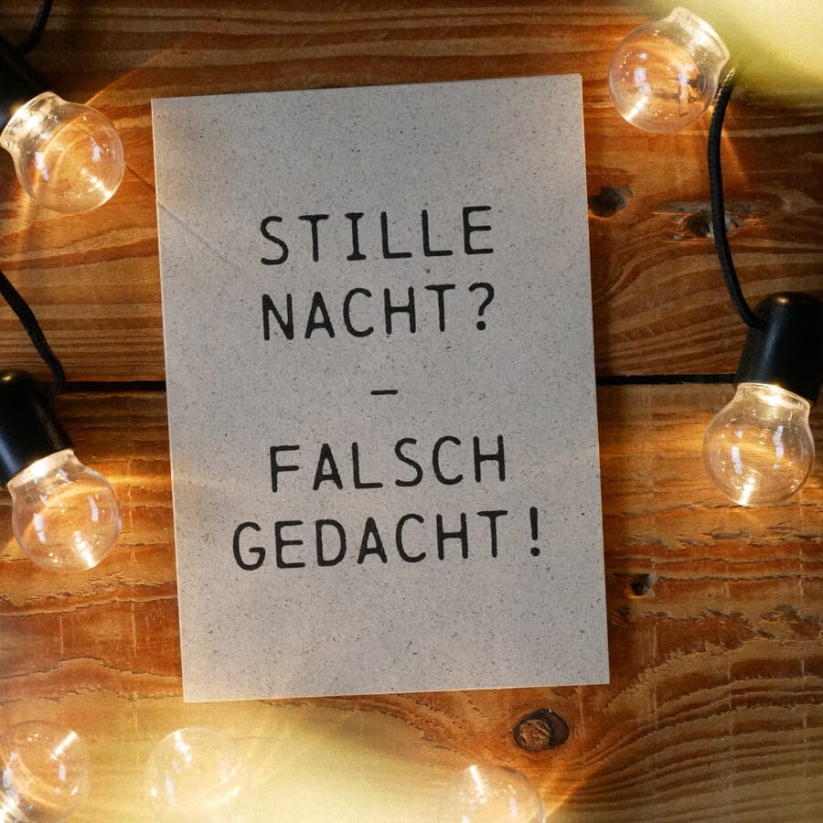 Karte mit der Aufschrift „Stille Nacht? – Falsch gedacht!“ auf einem Holztisch, umgeben von Glühbirnen.