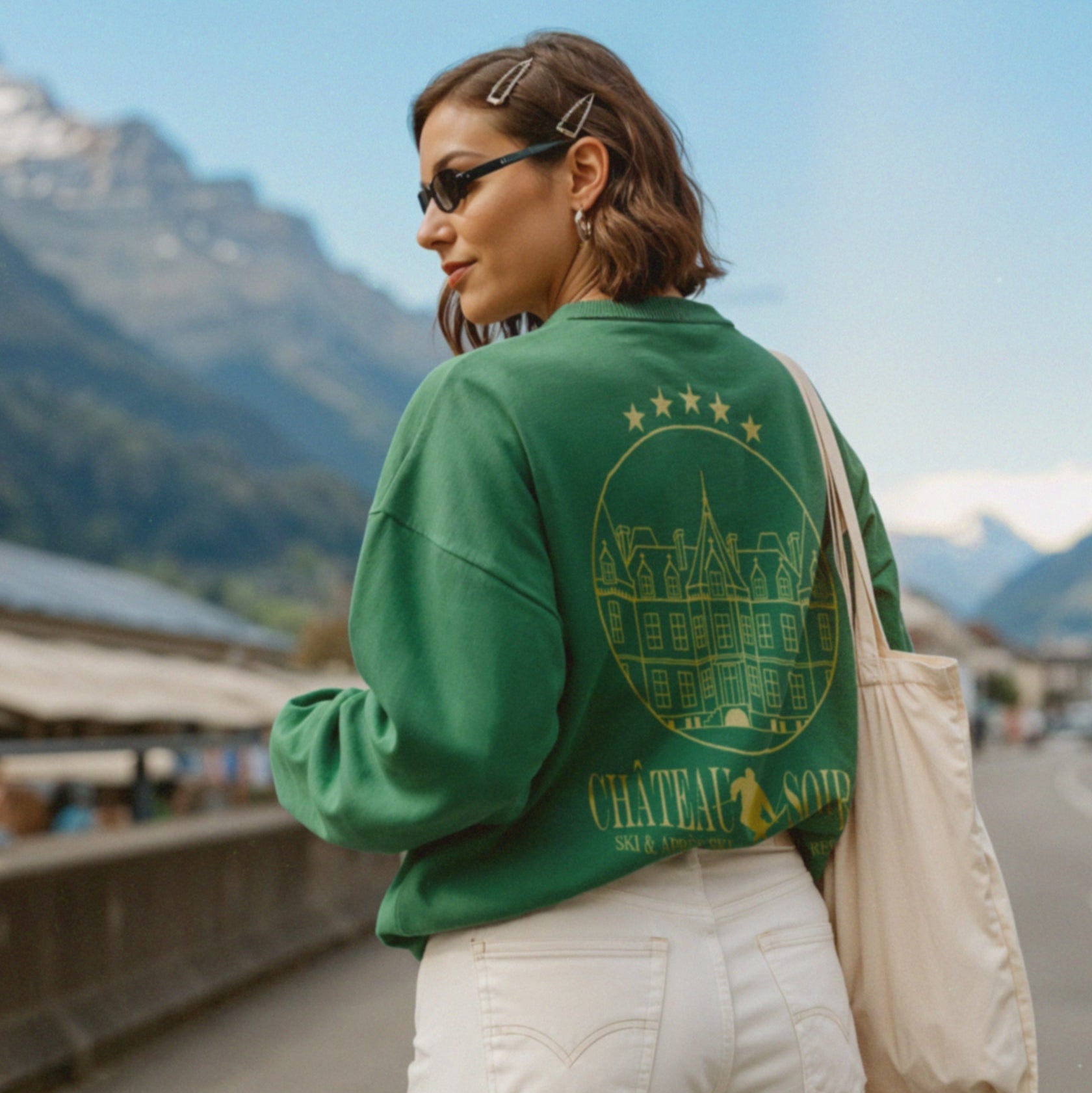 Eine Frau mit kurzen Haaren, Haarspangen und Sonnenbrille trägt einen grünen Pullover mit Château-Muster und eine weiße Hose. Sie trägt eine Tragetasche und steht im Freien mit Bergen und blauem Himmel im Hintergrund.