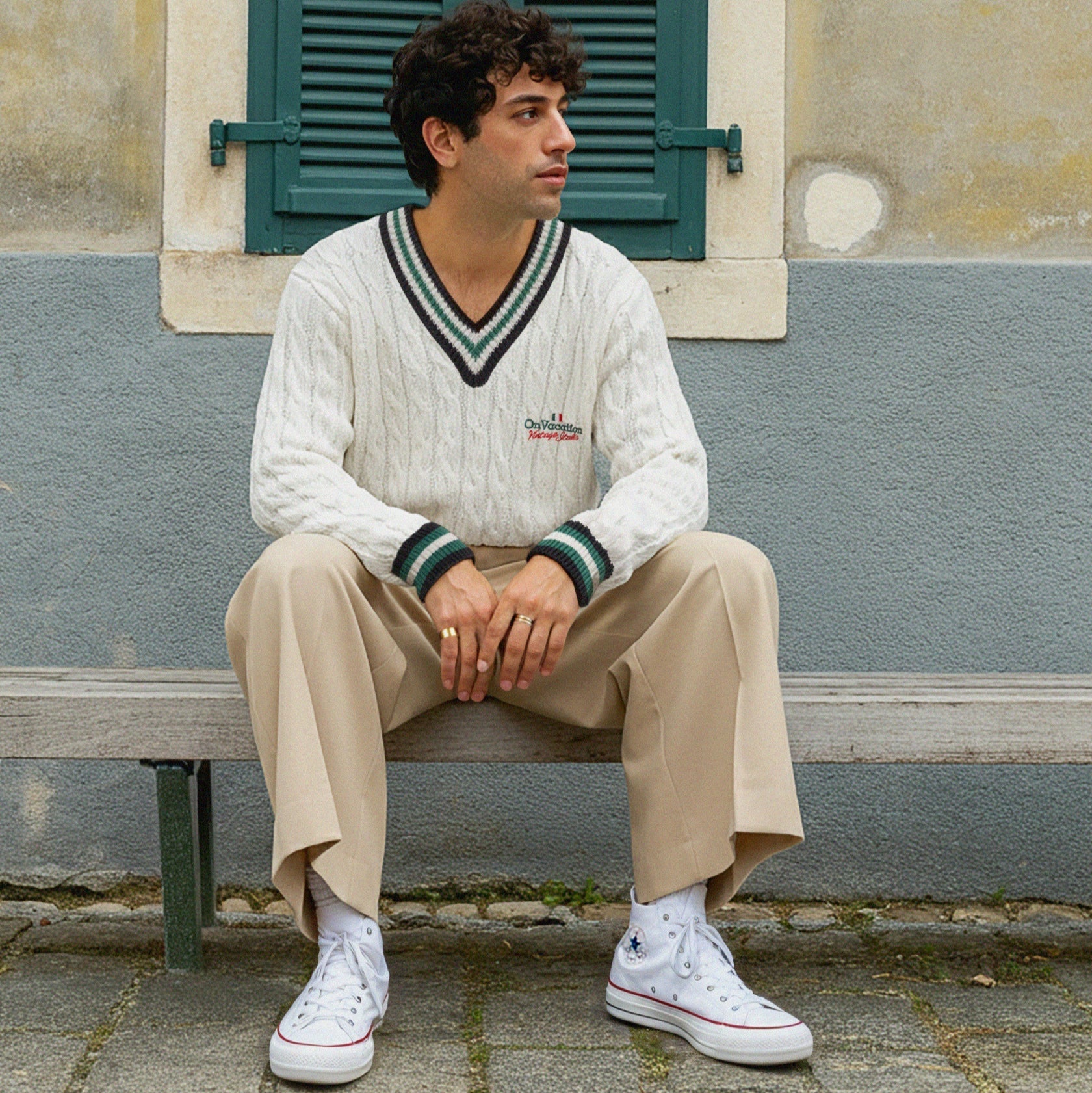 Ein Mann mit lockigem dunklem Haar sitzt auf einer Bank vor einer Wand mit einem grünen Fenster mit Fensterläden. Er trägt einen weißen Zopfstrickpullover, eine beigefarbene Hose und weiße High-Top-Turnschuhe und blickt zur Seite.