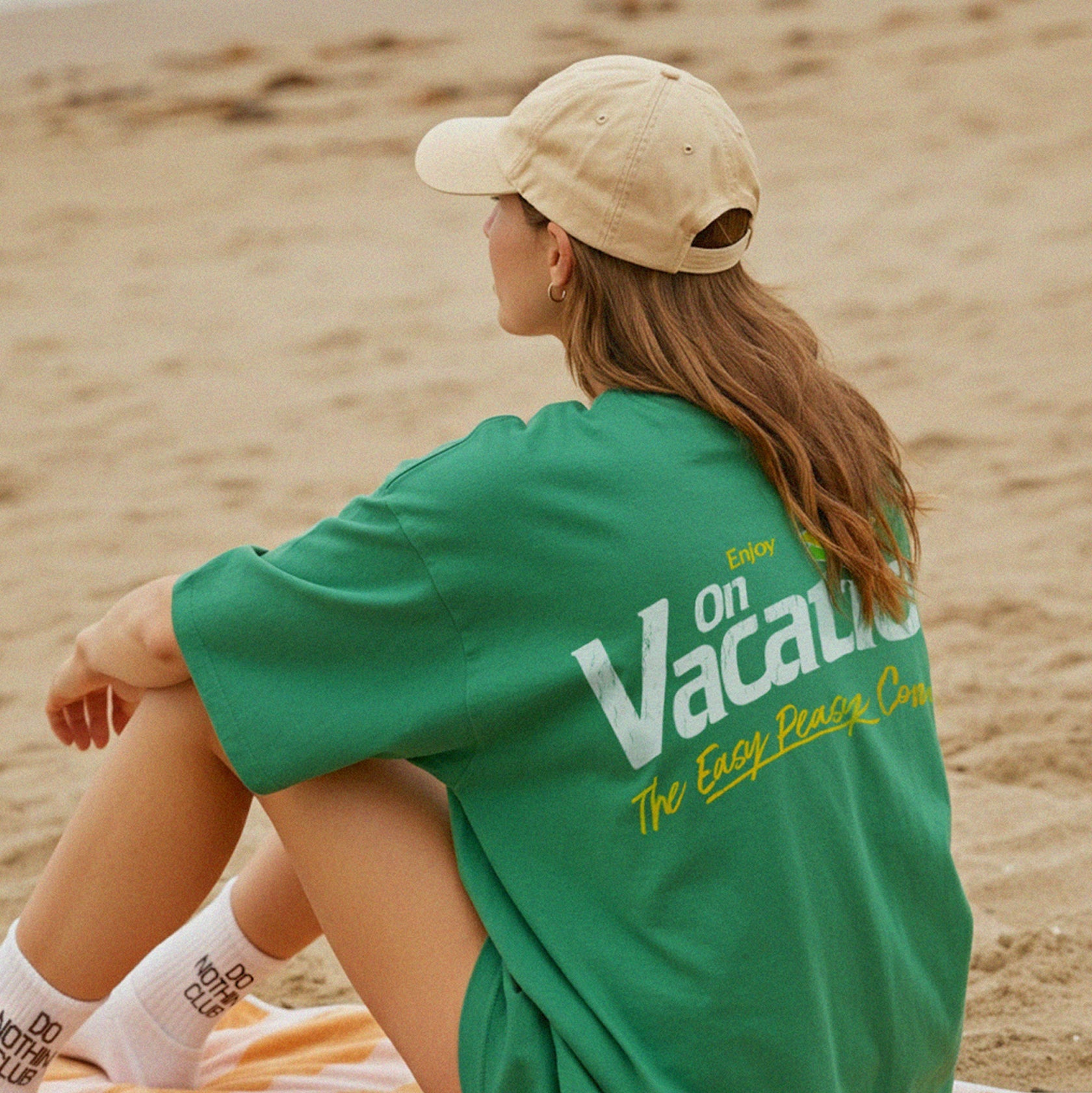 Eine Person mit einer beigen Mütze und einem grünen Hemd mit der Aufschrift Enjoy on Vacation sitzt auf einem gestreiften Handtuch am Strand und blickt auf das Meer. Sie trägt weiße Socken und scheint entspannt im Sand zu liegen.