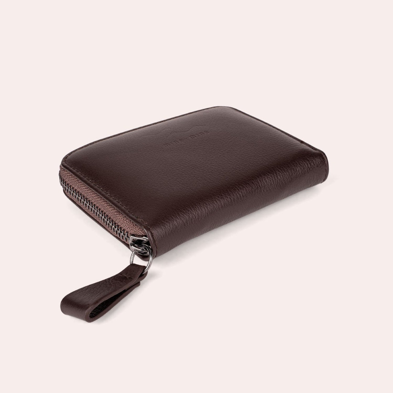 Geschlossenes, dunkelbraunes Lederportemonnaie mit Reißverschluss und Zuglasche auf hellbeigem Hintergrund.