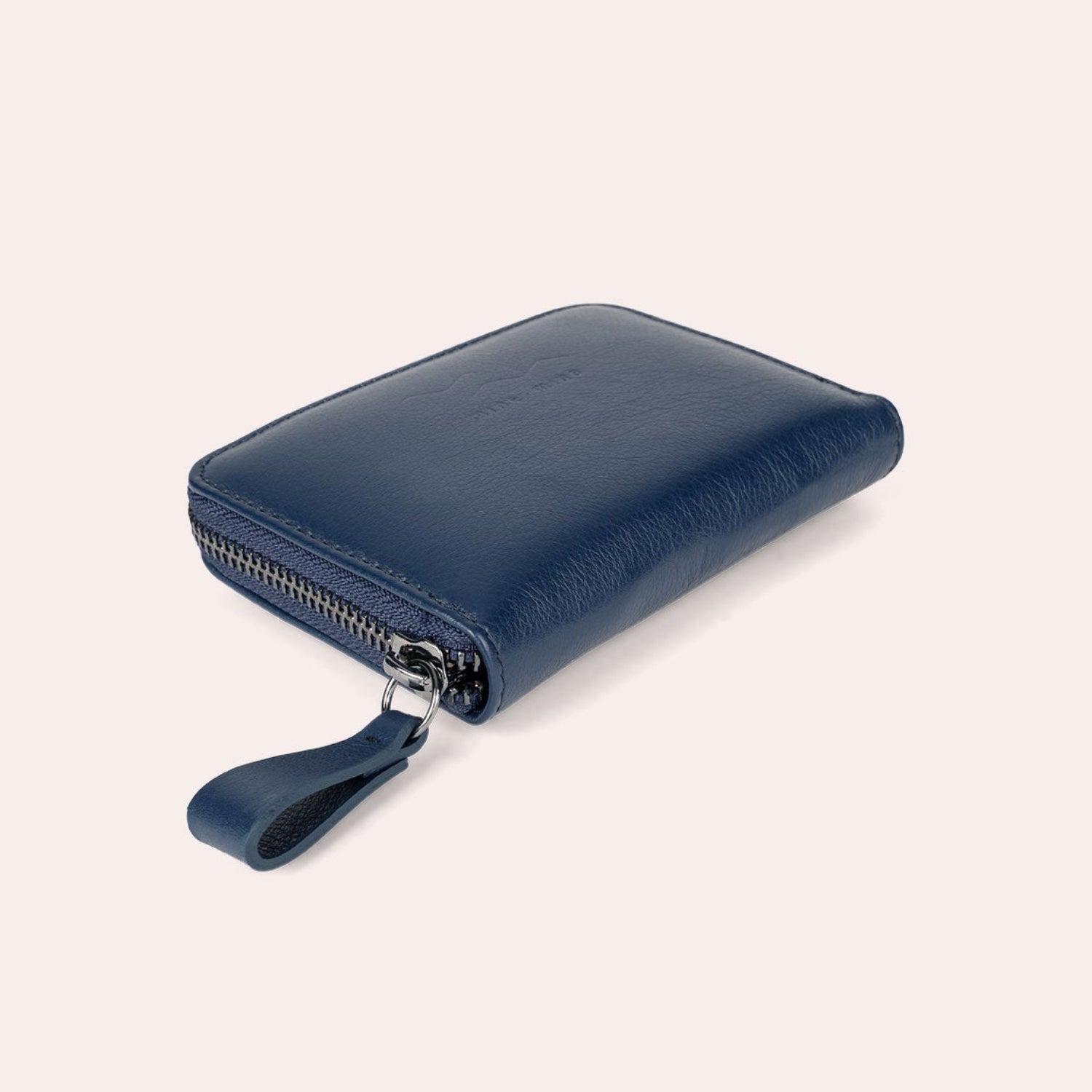 Ein blaues Lederportemonnaie mit Reißverschluss und passender Zuglasche, dargestellt auf einem hellbeigen Hintergrund. Die Brieftasche ist geschlossen und schräg positioniert, wodurch der Reißverschluss und das strukturierte Material hervorgehoben werden.