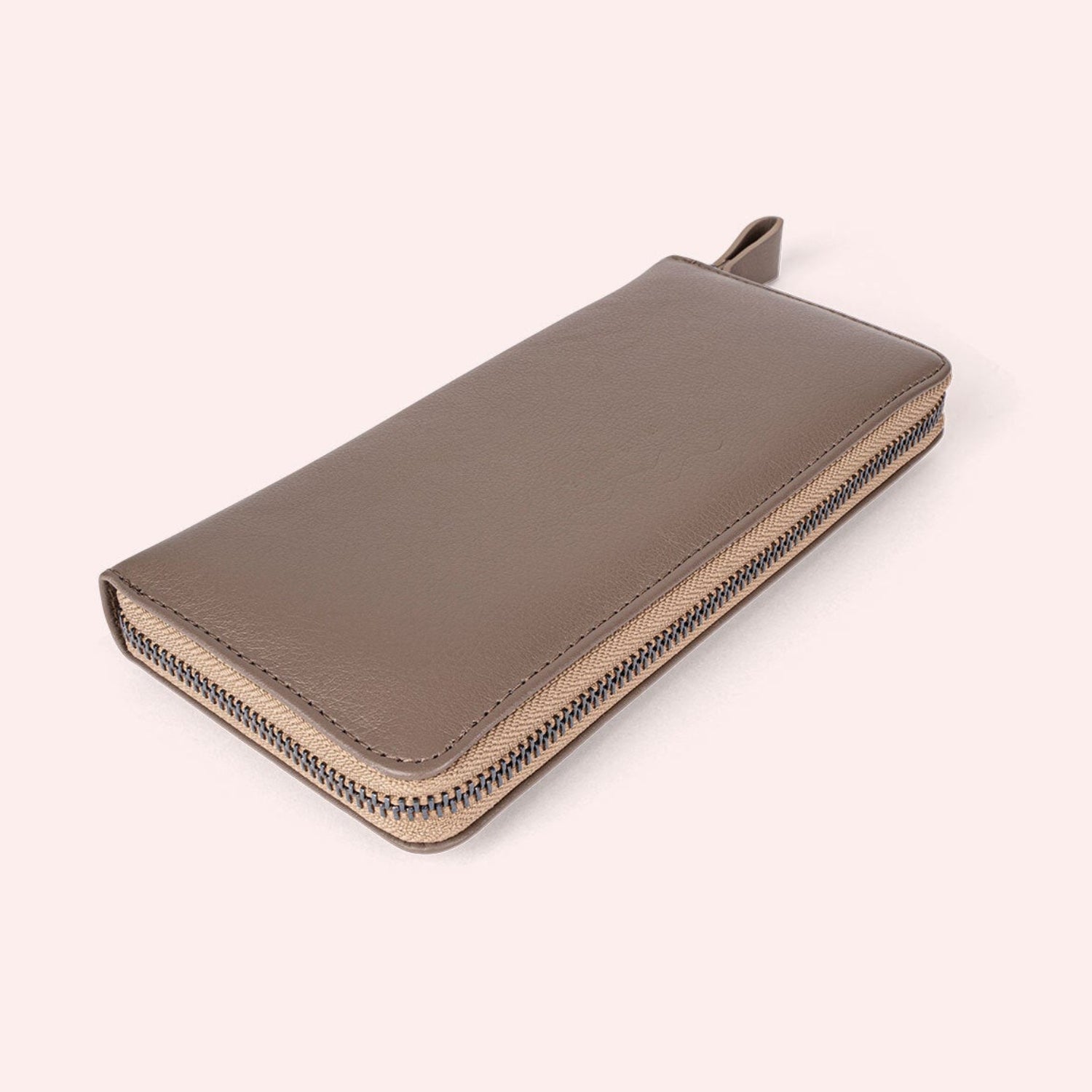 Ein taupefarbenes Lederportemonnaie mit Reißverschluss und einer Zuglasche auf hellrosa Hintergrund.