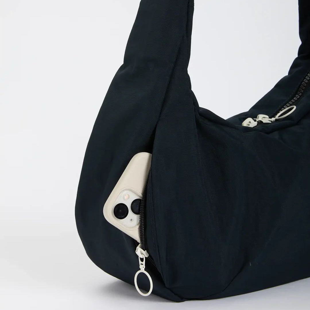 Eine schwarze Umhängetasche mit einer teilweise geöffneten Reißverschlusstasche, in der sich ein weißes Smartphone verbirgt. Die Tasche ist vor einem schlichten weißen Hintergrund platziert.