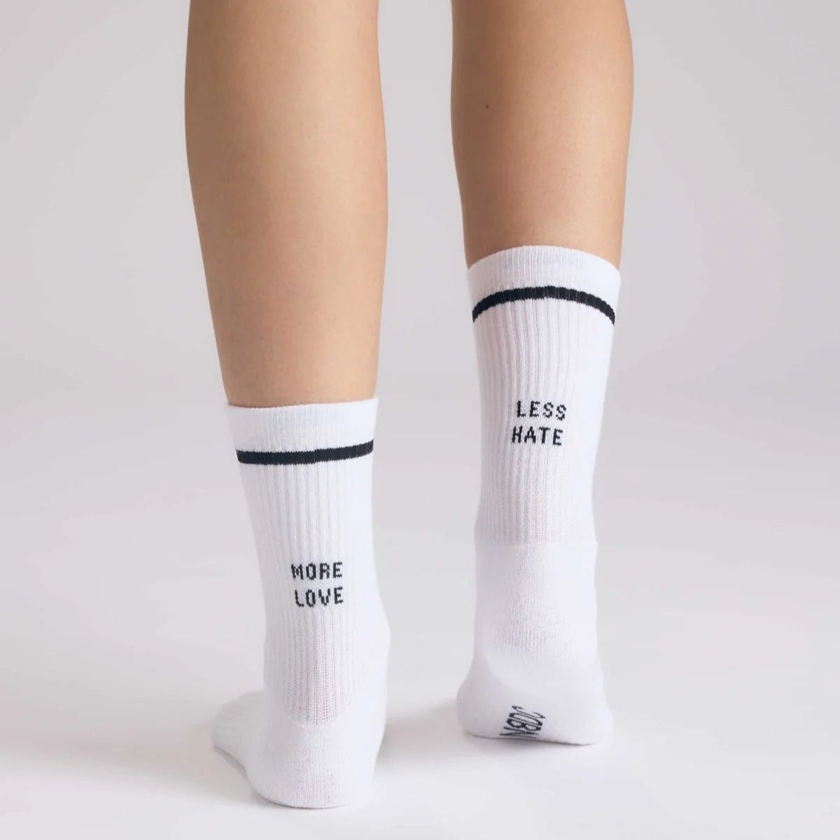 Eine Person steht auf einer weißen Fläche und trägt weiße Socken mit schwarzen Streifen; auf der linken Socke steht MEHR LIEBE und auf der rechten Socke steht WENIGER HASS auf der Rückseite.