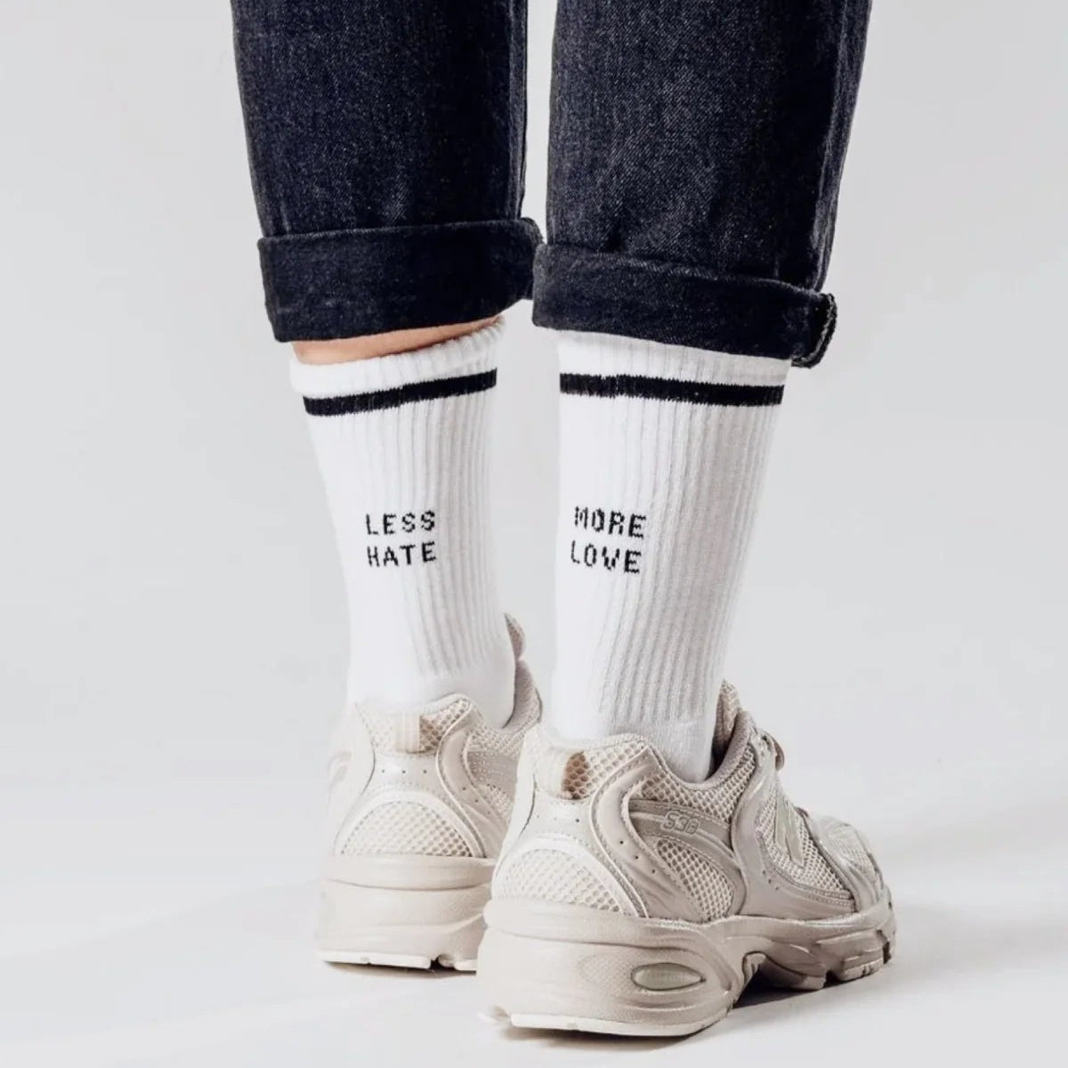 Person trägt weiße Turnschuhe, dunkle Jeans mit Bündchen und weiße Socken. Auf einer Socke steht LESS HATE und auf der anderen MORE LOVE in schwarzen Buchstaben. Die Person steht auf einer hellen Fläche.