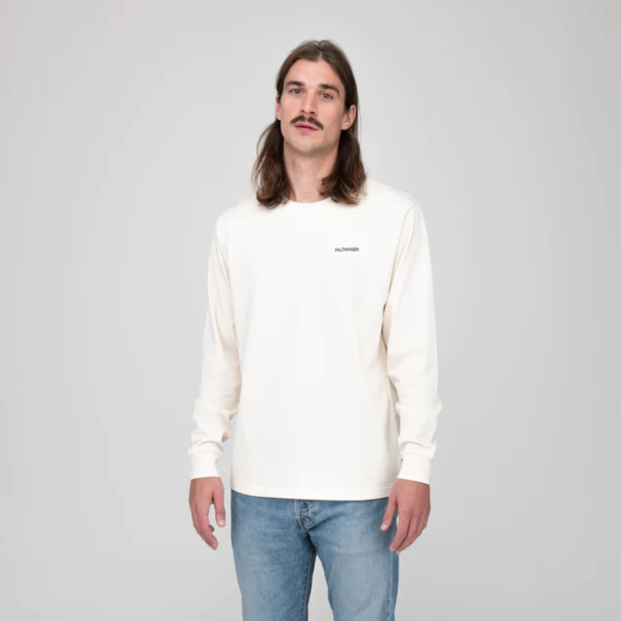 Ein Mann mit langen Haaren und Schnurrbart trägt ein weißes Langarmshirt und blaue Jeans vor grauem Hintergrund.