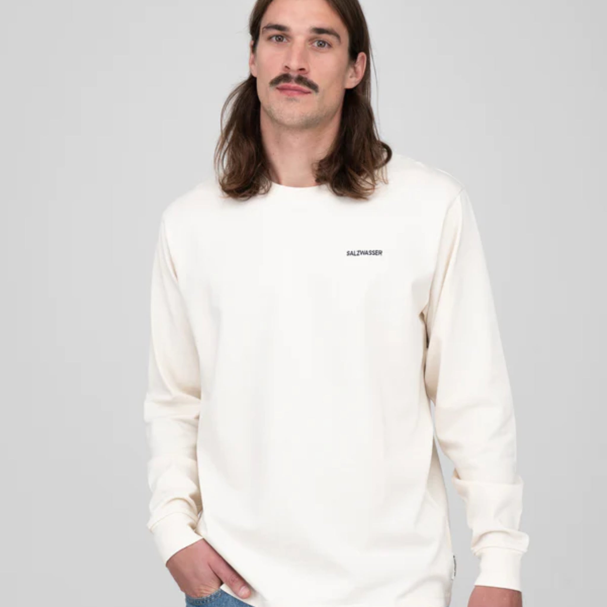 Mann mit langen Haaren und Schnurrbart trägt ein weißes Langarmshirt mit Aufdruck "SALZWASSER" vor grauem Hintergrund.