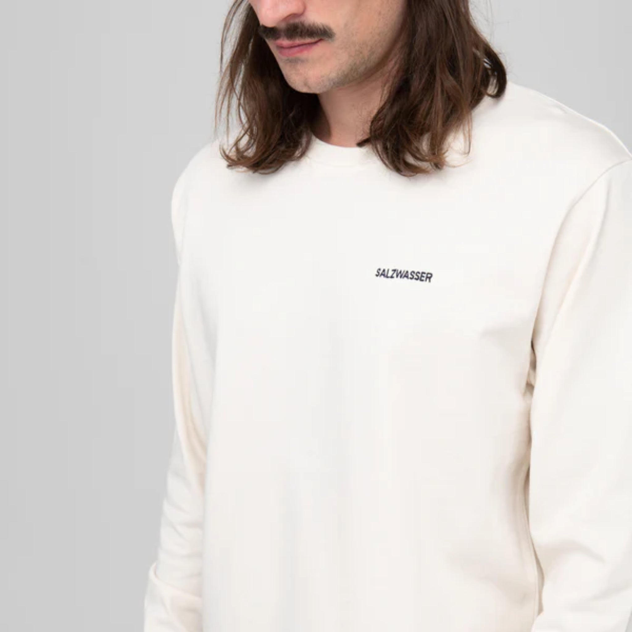 Mann mit langen Haaren trägt ein cremefarbenes Sweatshirt mit dem Aufdruck "SALZWASSER".
