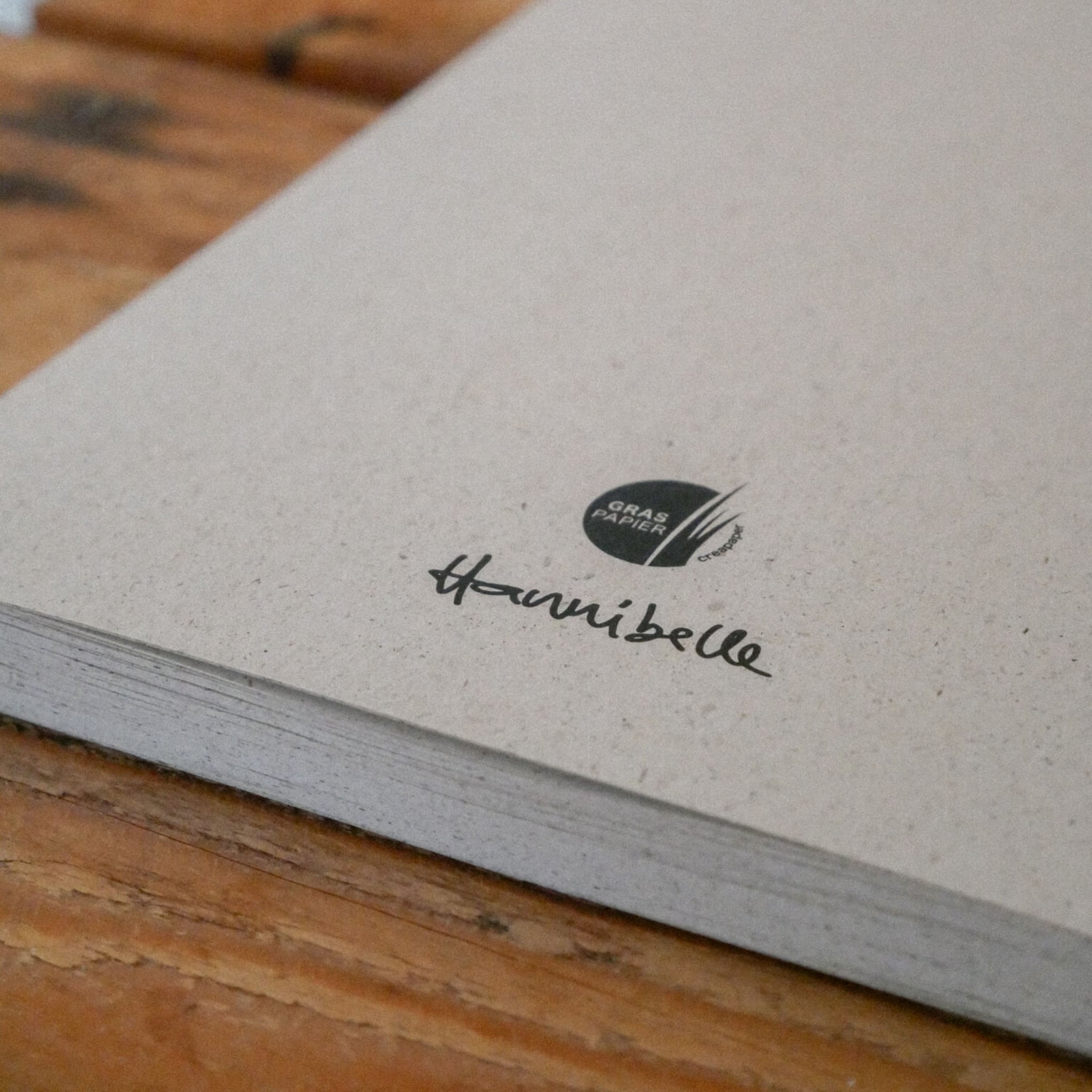 Eine Nahaufnahme eines grauen, strukturierten Notizbuchs, das auf einer hölzernen Oberfläche liegt. Auf dem Einband befindet sich ein kleines schwarzes Logo mit der Aufschrift GRAP PAPER und darunter eine handschriftliche Unterschrift mit der Aufschrift hannibelle.