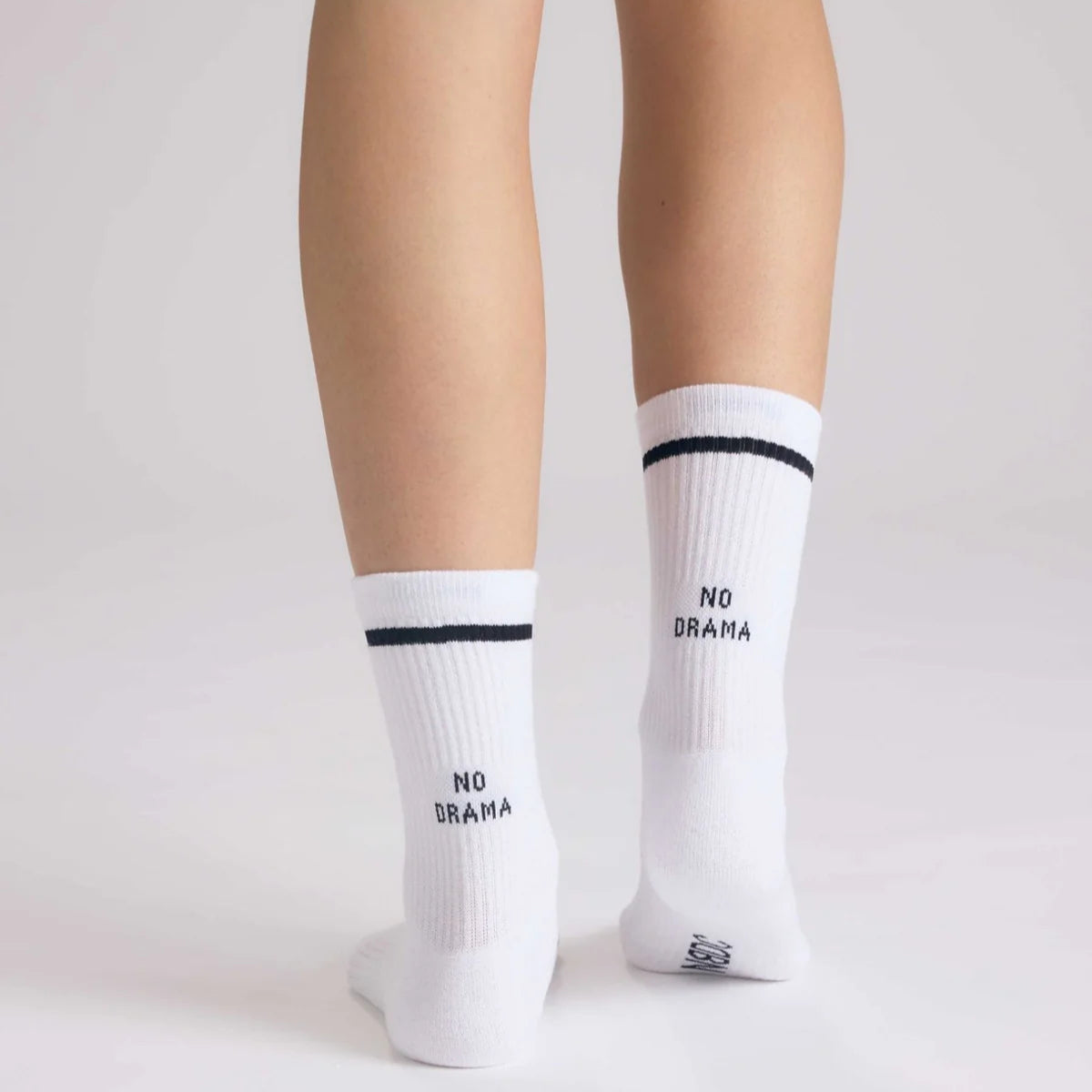Eine Person steht auf einer hellen Fläche und trägt weiße Socken mit einem schwarzen Streifen und der Aufschrift NO DRAMA auf der Rückseite jeder Socke.