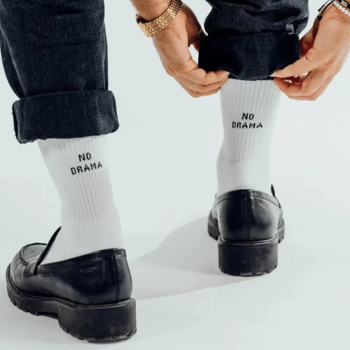 Eine Person, die eine schwarze Hose, schwarze Halbschuhe und weiße Socken mit der Aufschrift NO DRAMA trägt, passt die Manschetten ihrer Hose an. Die Person trägt auch eine Uhr und ein Armband.