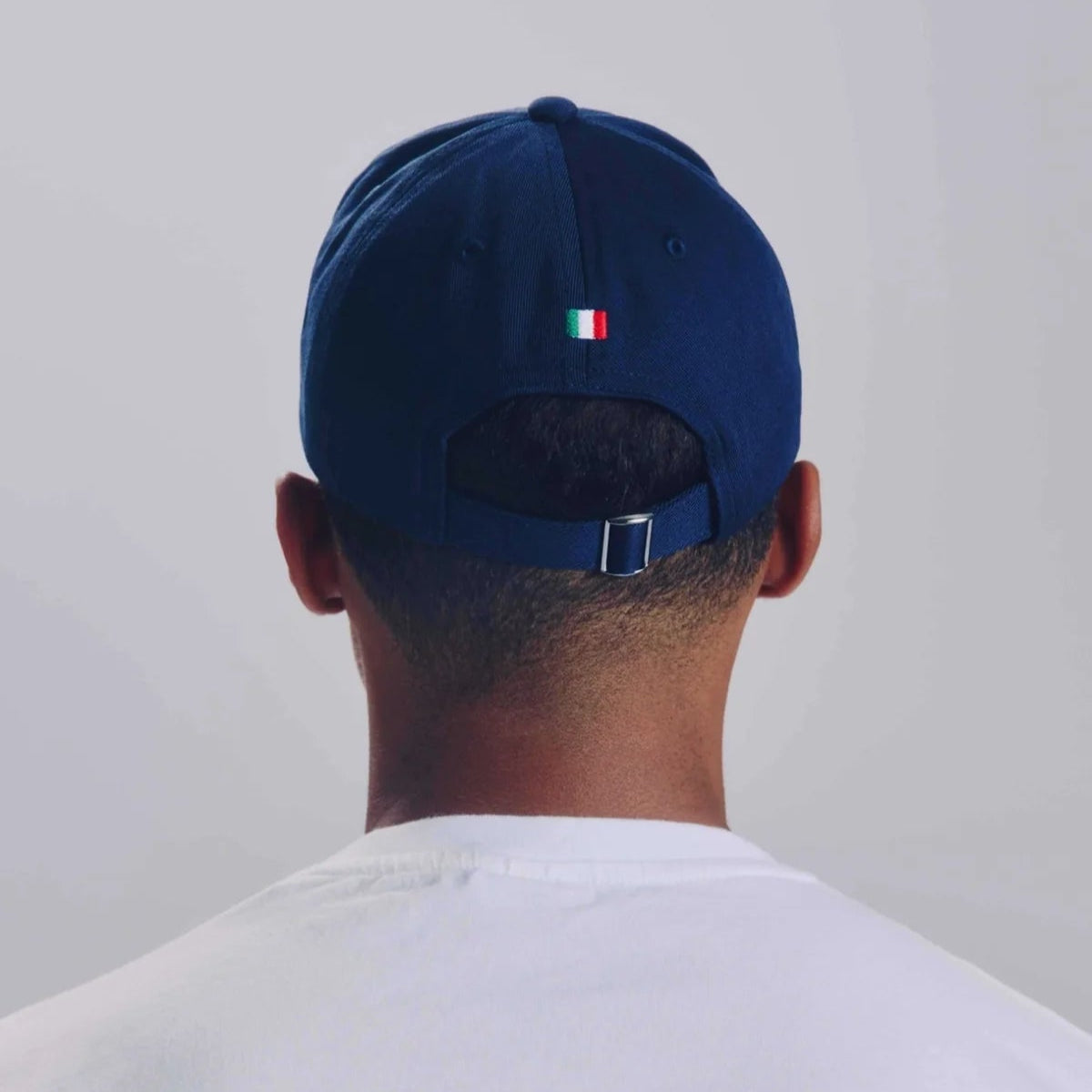 Eine Person, die ein weißes Hemd und eine marineblaue Baseballmütze trägt, wird von hinten gezeigt. Die Kappe hat ein kleines italienisches Flaggendetail über dem verstellbaren Riemen. Der Hintergrund ist einfarbig und in hellen Tönen gehalten.