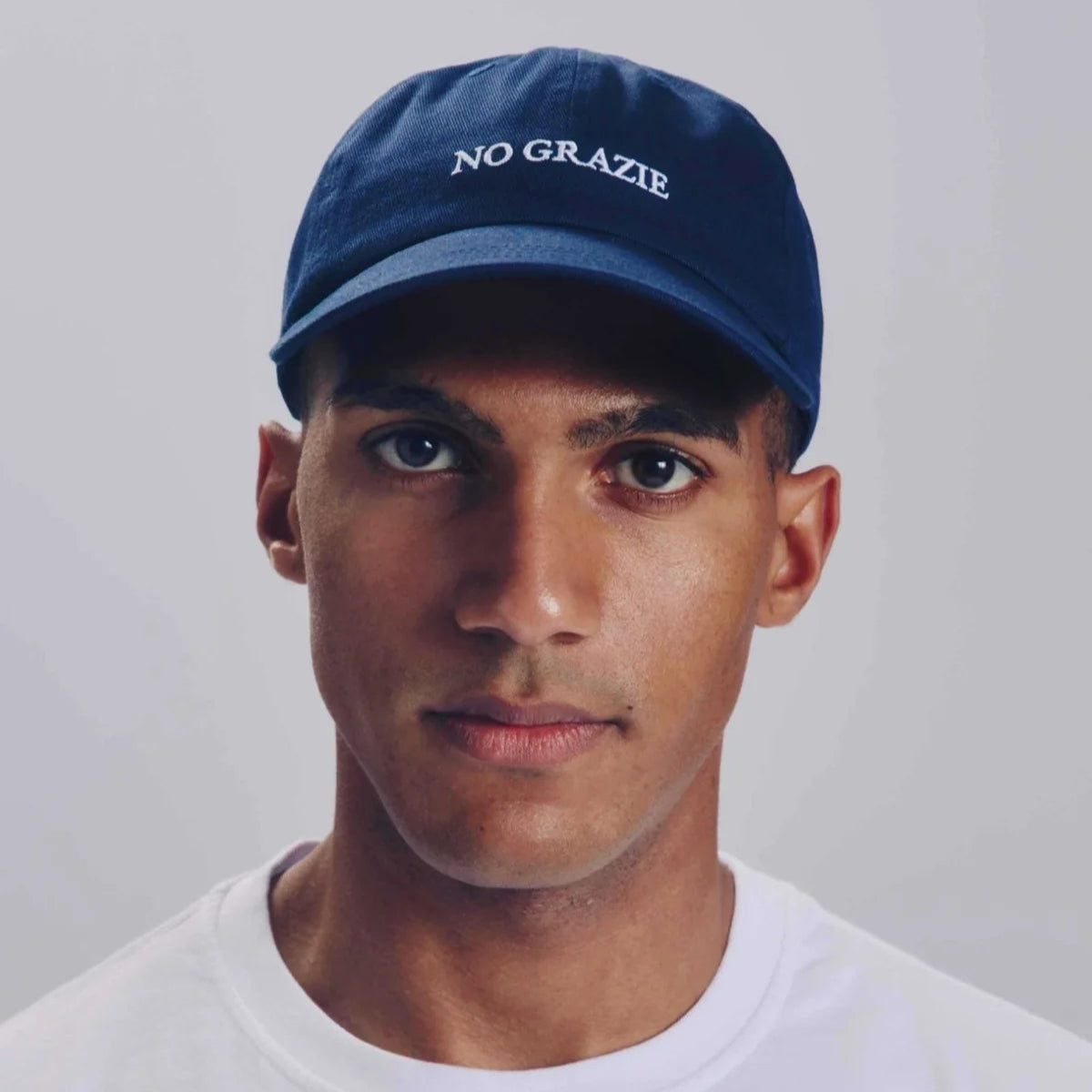 Eine Person, die eine marineblaue Baseballkappe mit der weiß aufgestickten Aufschrift NO GRAZIE und ein schlichtes weißes T-Shirt trägt und vor einem neutralen Hintergrund direkt in die Kamera schaut.