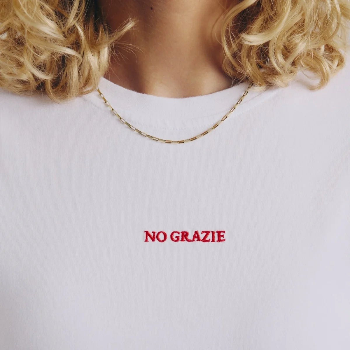 Nahaufnahme einer Person, die ein weißes Hemd mit der Aufschrift NO GRAZIE in roter Schrift auf der Brust trägt. Die Person hat lockiges blondes Haar und trägt eine dünne goldene Halskette.