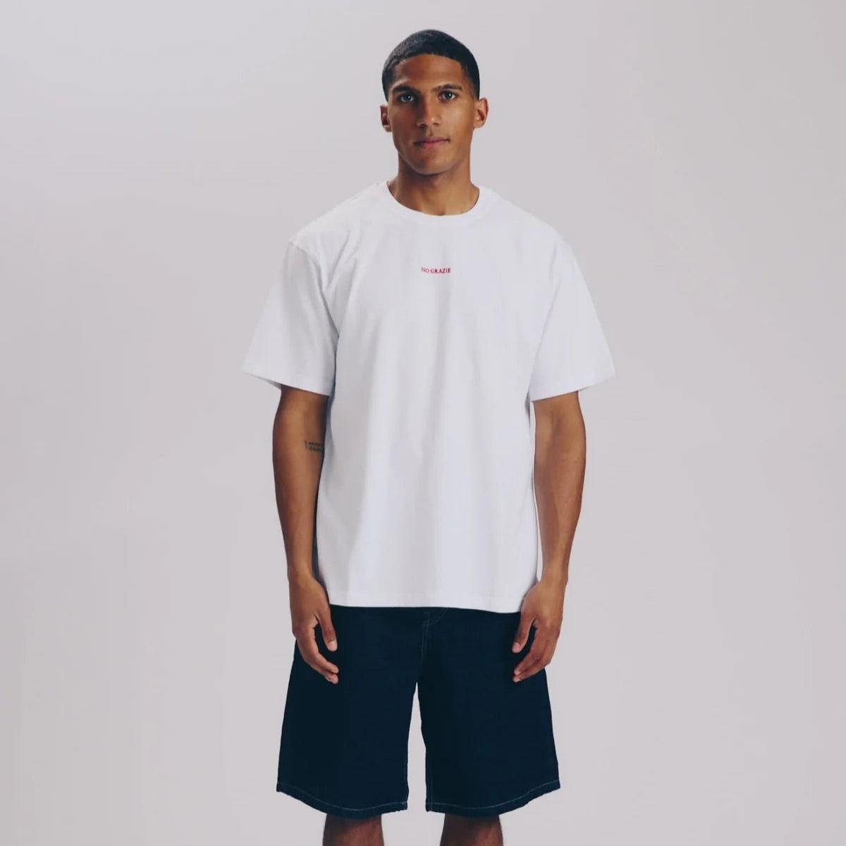 Ein Mann steht mit dem Gesicht nach vorne und trägt ein schlichtes weißes T-Shirt, dunkle knielange Shorts, weiße Socken und weiße Turnschuhe vor einem hellgrauen Hintergrund.