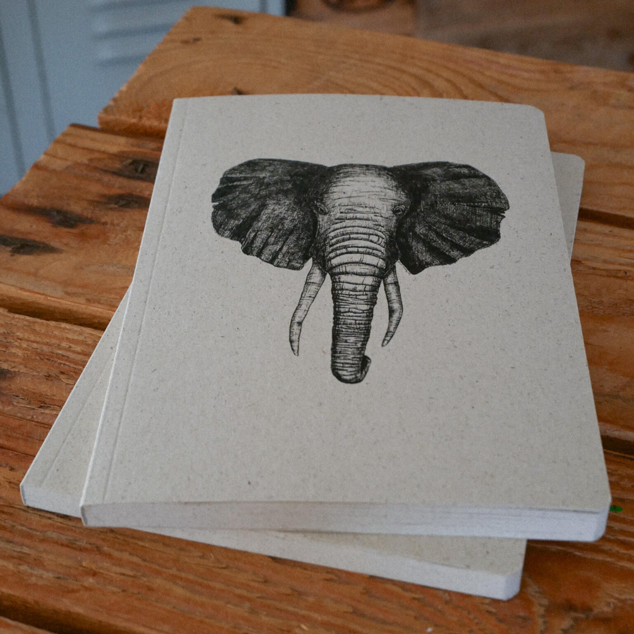 Zwei beigefarbene Notizbücher mit einer detaillierten schwarzen Skizze eines Elefantenkopfes auf dem Einband, die auf einer rustikalen Holzunterlage liegen.