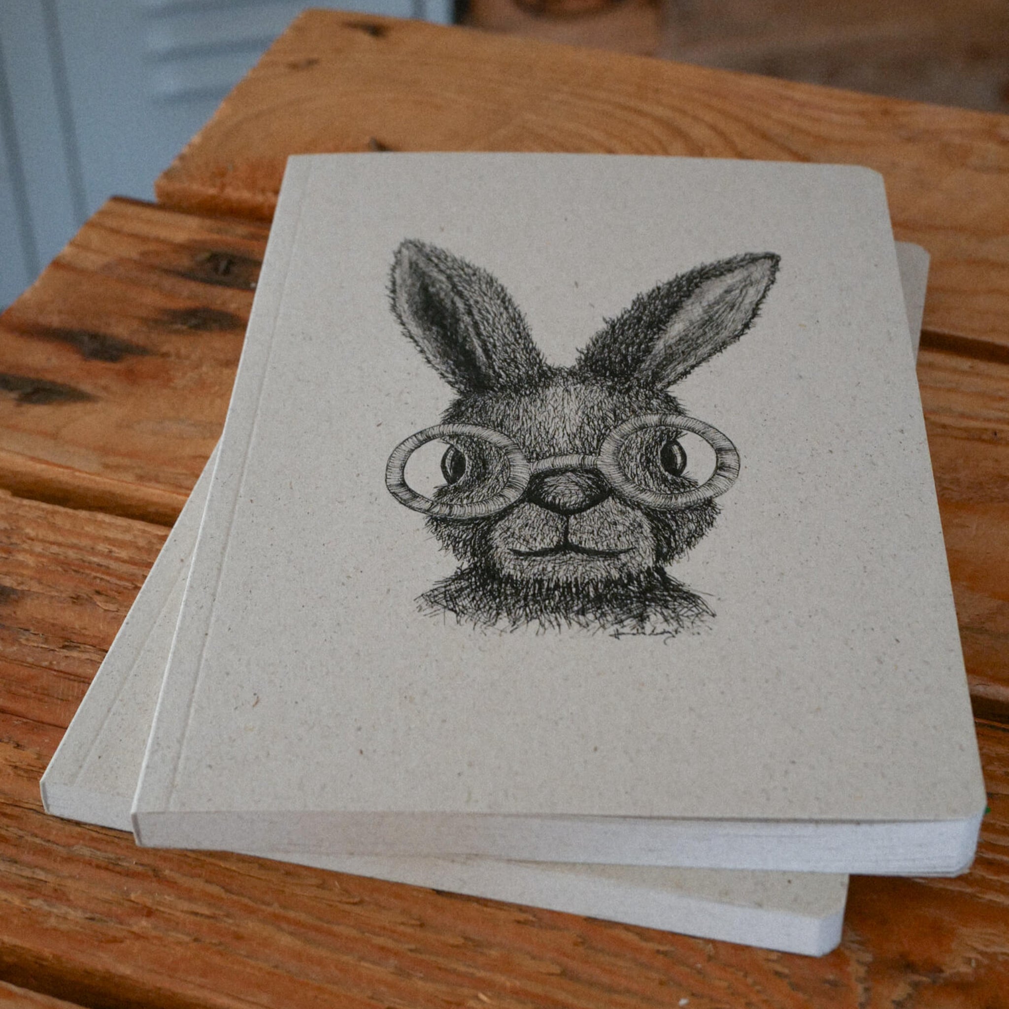 Ein Notizbuch mit einer schwarzen Tuschezeichnung eines Kaninchens mit runder Brille auf dem Einband, das auf einer hölzernen Unterlage ruht, unter der ein weiteres Notizbuch teilweise sichtbar ist.