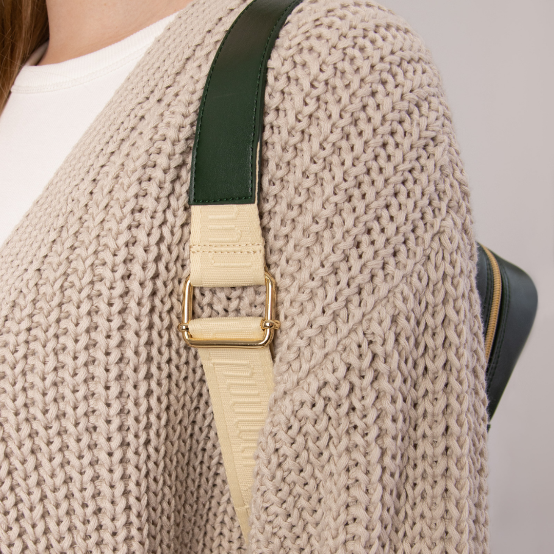 Nahaufnahme eines beigen, grob gestrickten Cardigans und einem grünen Taschengurt über der Schulter einer Person.