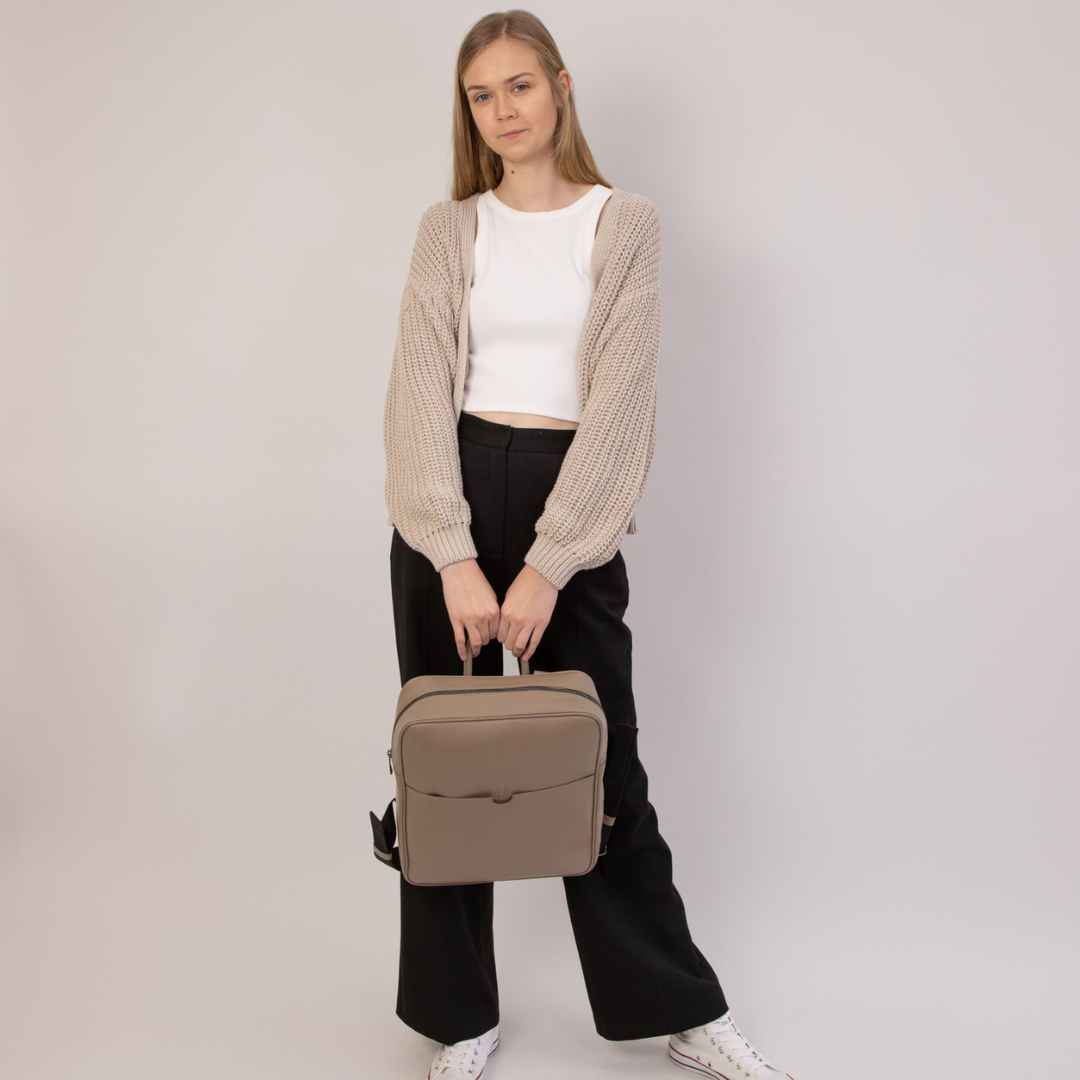 Frau in beiger Strickjacke und schwarzer Hose hält eine beige Tasche vor einem neutralen Hintergrund.