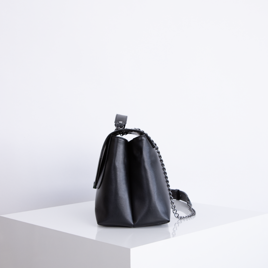 Schwarze Lederhandtasche mit Kettengriff steht auf einem weißen Podest vor hellem Hintergrund.