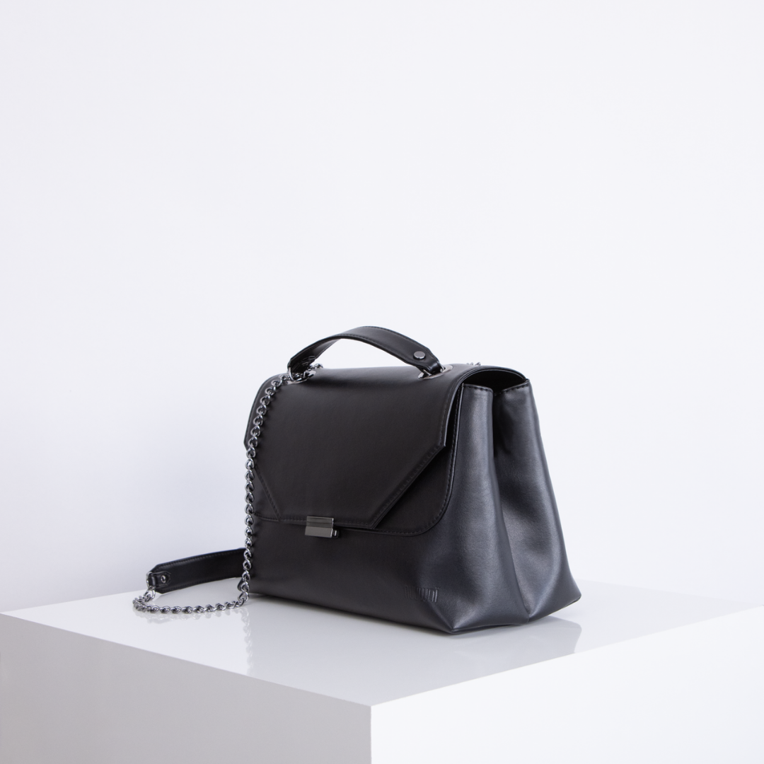 Schwarze Ledertasche mit Kettengurt auf einem weißen Sockel vor weißem Hintergrund.