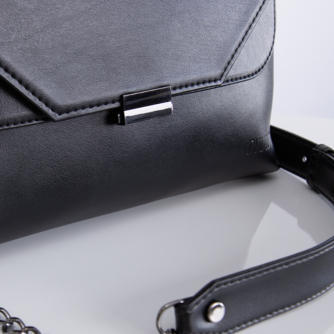 Schwarze Ledertasche mit silbernem Verschluss liegt auf weißer Oberfläche, eine Kette und ein Riemen sind sichtbar.