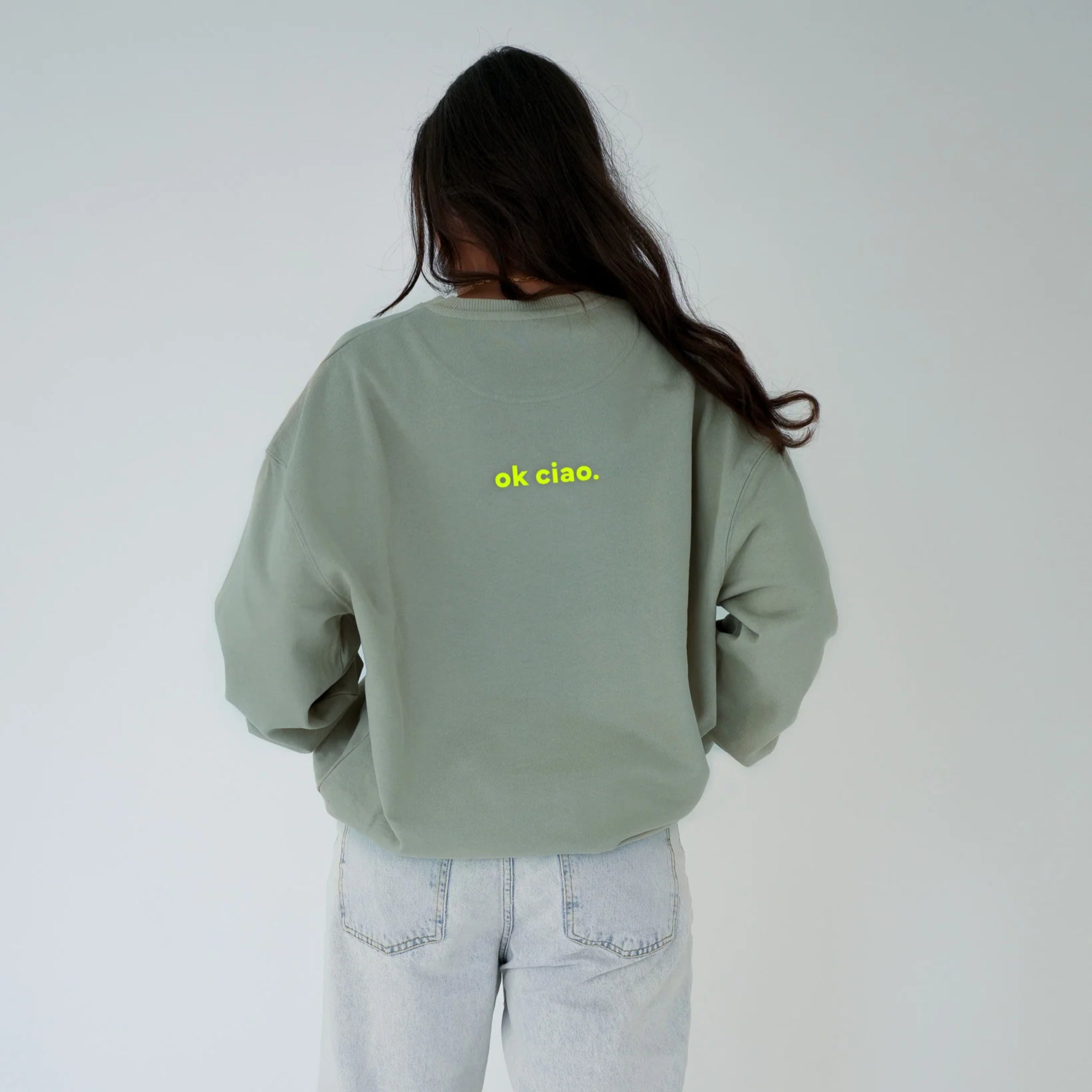 Eine Person mit langen braunen Haaren, von hinten gesehen, trägt ein hellgrünes Sweatshirt mit der gelben Aufschrift ok ciao. auf dem Rücken und eine helle Jeans, die vor einem schlichten hellen Hintergrund steht.