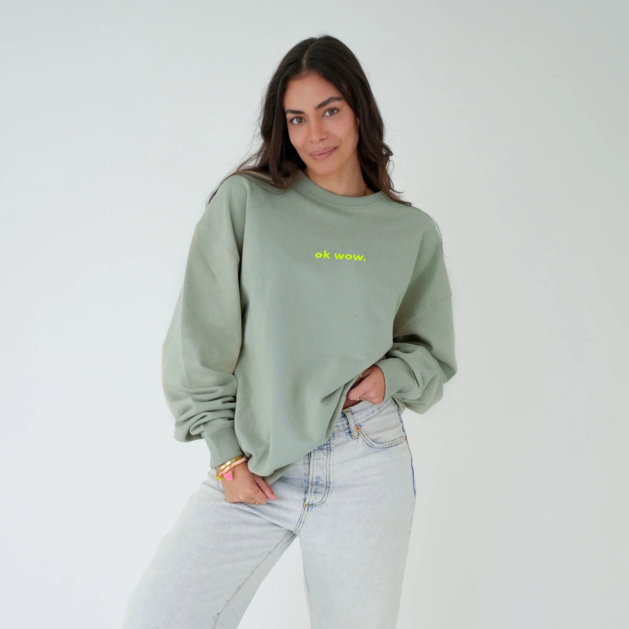 Eine Frau mit langen dunklen Haaren trägt ein lockeres salbeigrünes Sweatshirt mit der Aufschrift ok wow. auf der Vorderseite, hellblaue Jeans, und steht vor einem schlichten weißen Hintergrund, eine Hand in der Tasche.