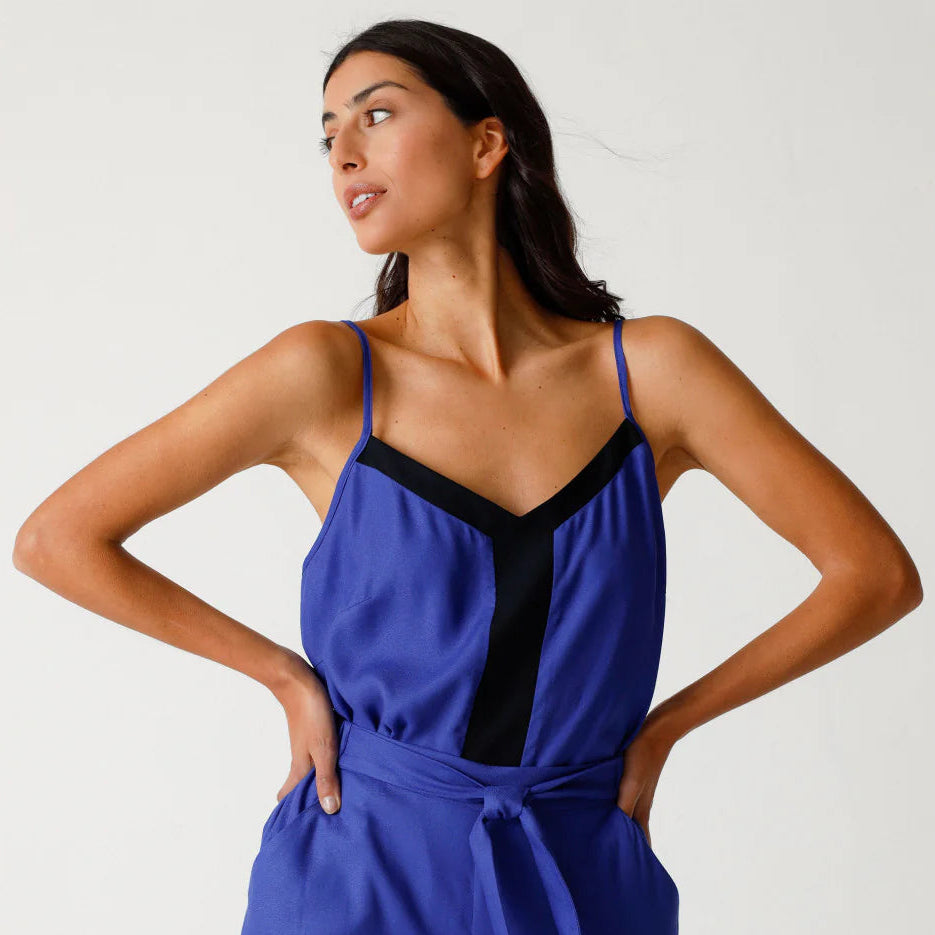 Eine Frau steht mit den Händen in den Hüften und trägt einen ärmellosen blauen Overall mit dünnen Trägern, einem V-förmigen schwarzen Ausschnitt auf dem Mieder und einer Hose mit weitem Bein. Sie blickt selbstbewusst zur Seite vor einem schlichten Hintergrund.