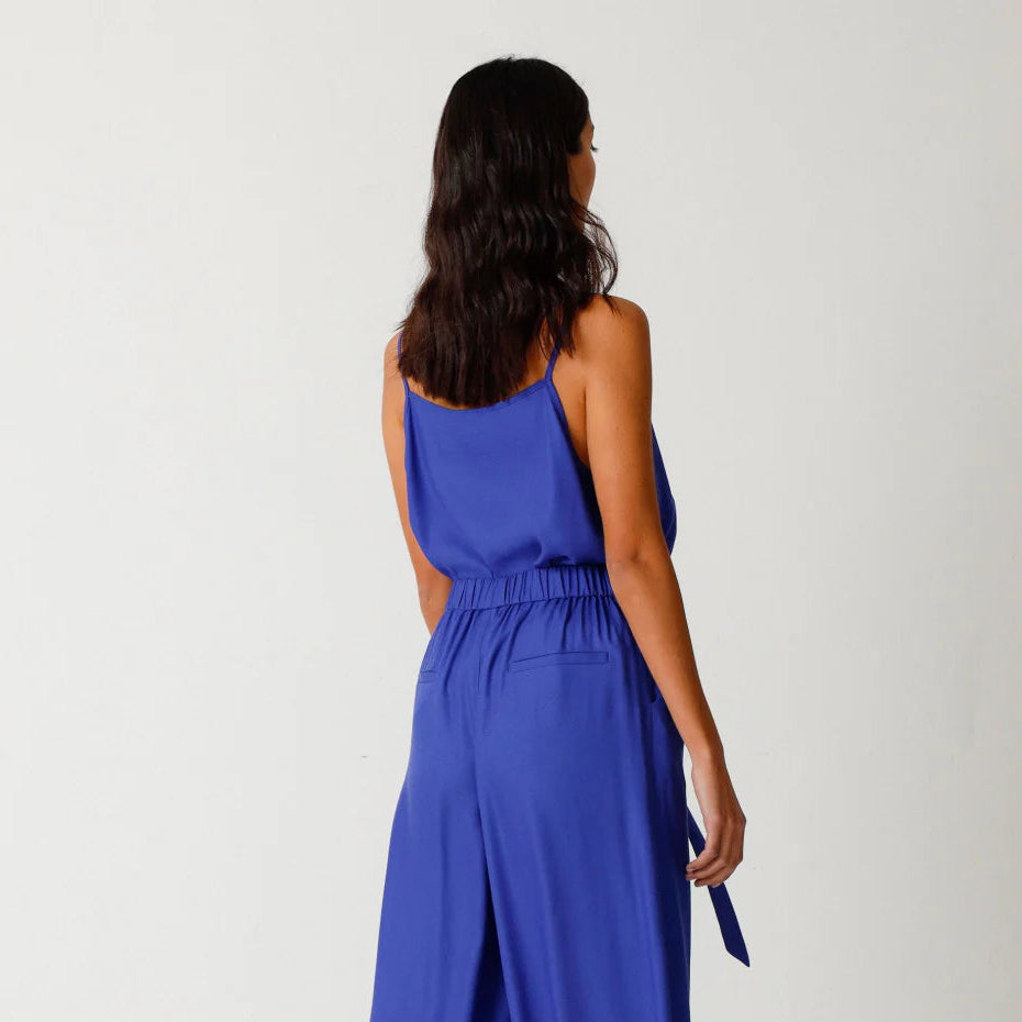 Eine Frau steht mit dem Gesicht zur Wand und trägt einen ärmellosen, königsblauen Overall mit hochgeschlossener Taille und einer Hose mit weitem Bein, dazu schwarze, offene Sandalen. Der Hintergrund ist einfarbig weiß.