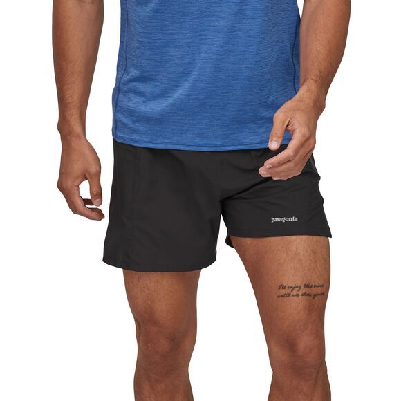 Mann trägt ein blaues T-Shirt und schwarze Shorts mit einem "Patagonia"-Logo; ein Tattoo ist auf seinem Oberschenkel sichtbar.