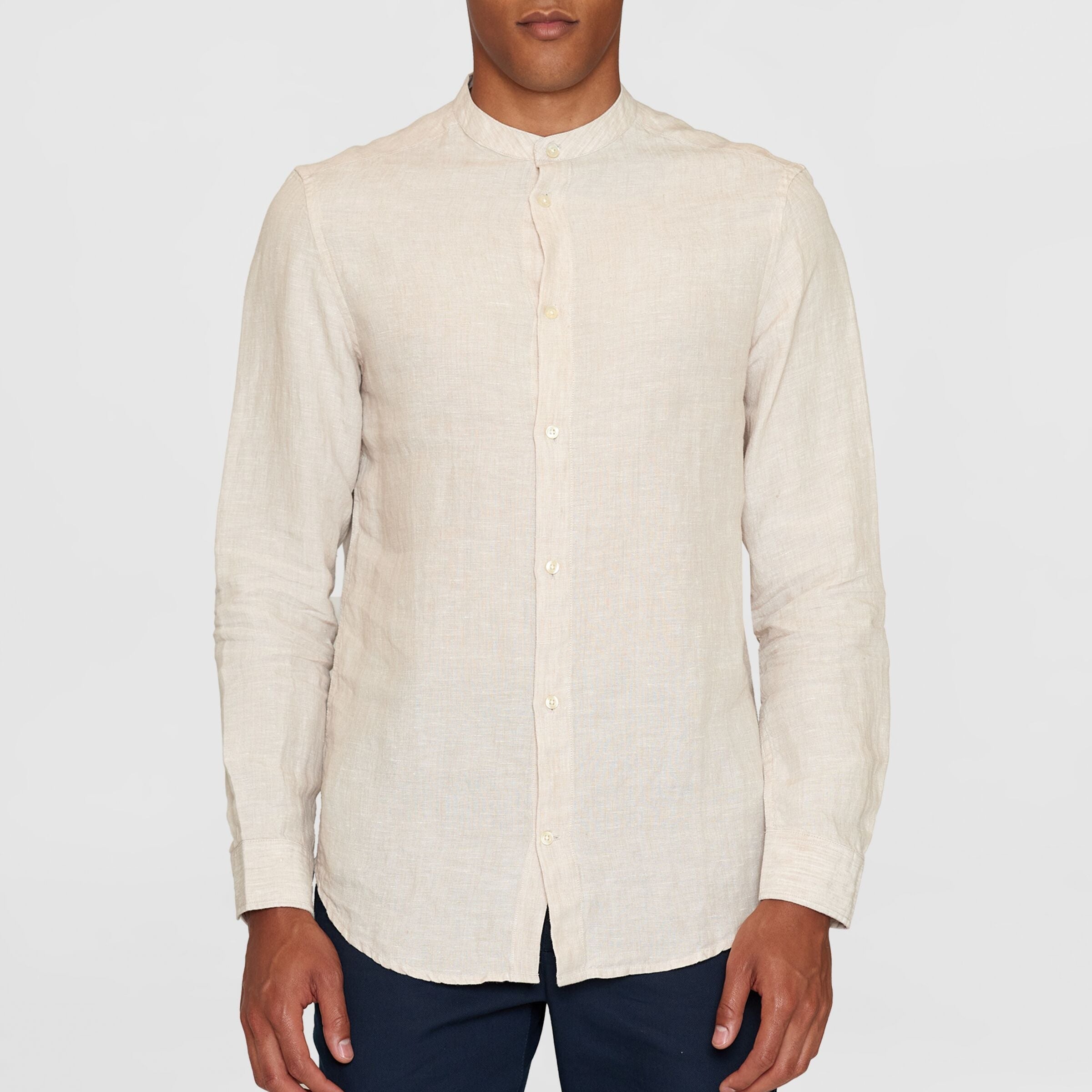 Mann trägt ein helles Leinenhemd mit Stehkragen und dunkelblauer Hose vor grauem Hintergrund.