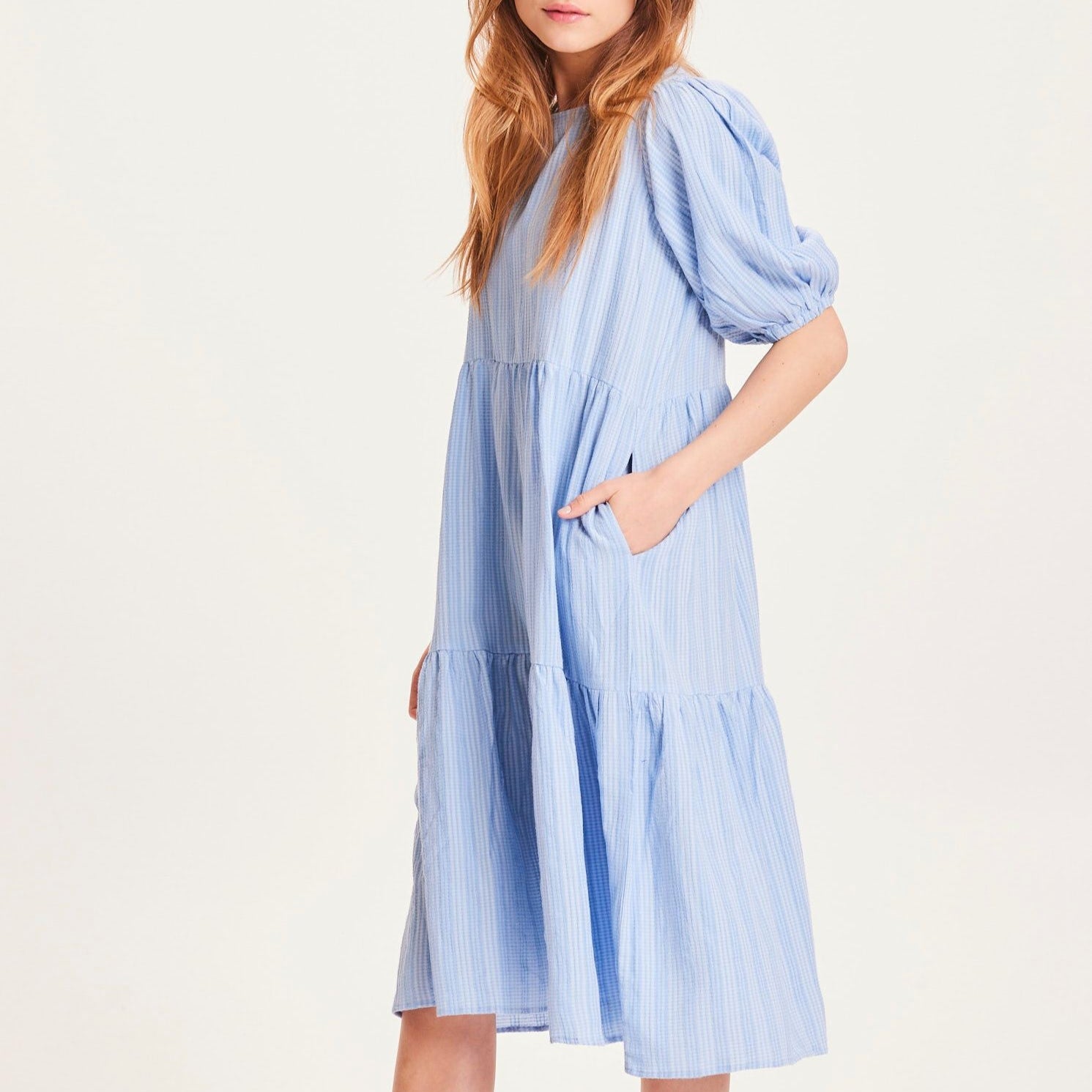Person in a light blue, striped dress with short, puffed sleeves, and hands in the pockets, against a plain background.