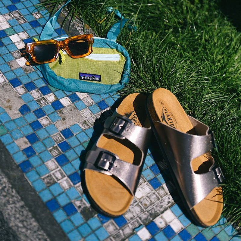 Sonnenbrille und Bauchtasche auf blauer Mosaikfliese neben einer Wiese mit silbernen Sandalen.