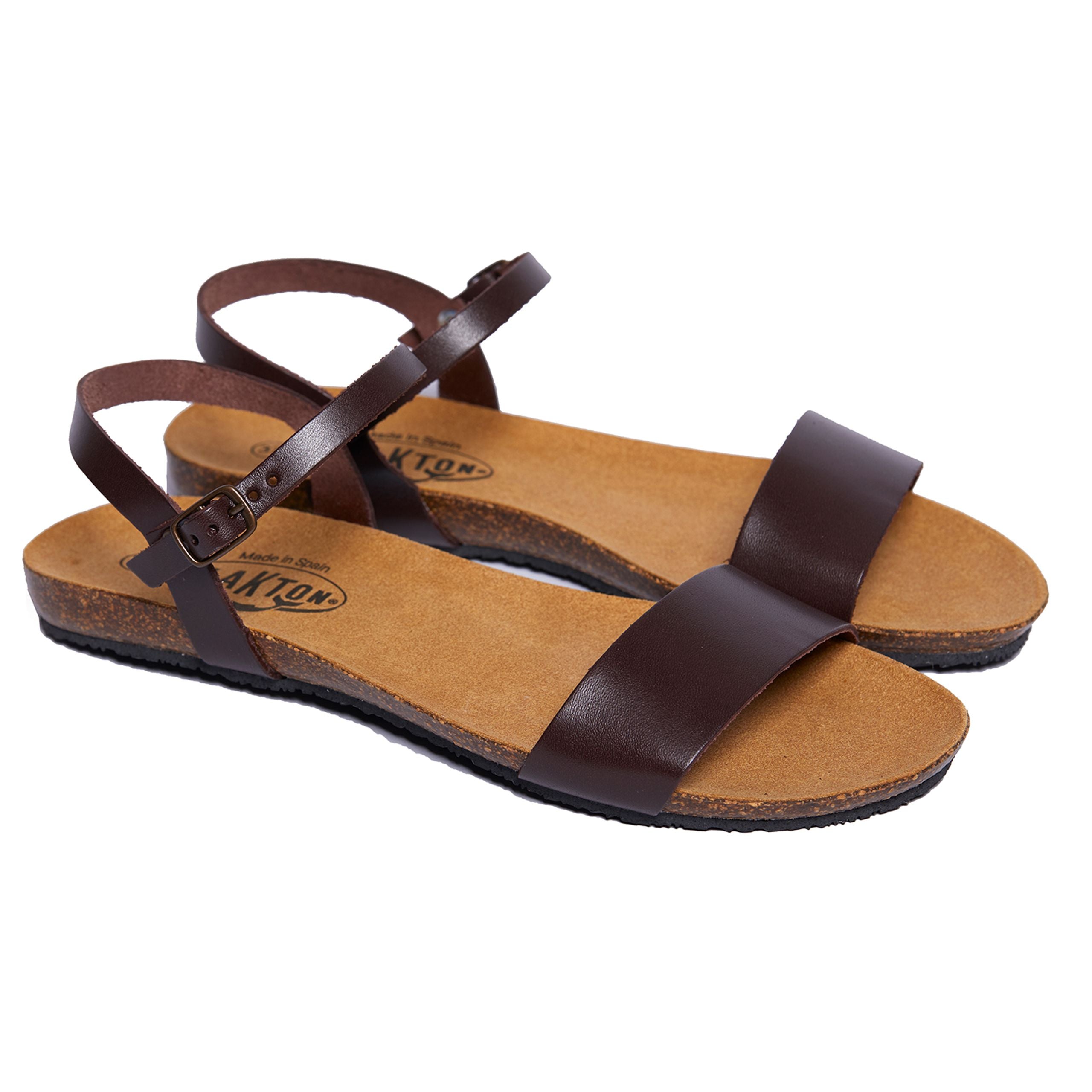 Braune Ledersandalen mit Knöchelriemen und flacher Sohle auf weißem Hintergrund.