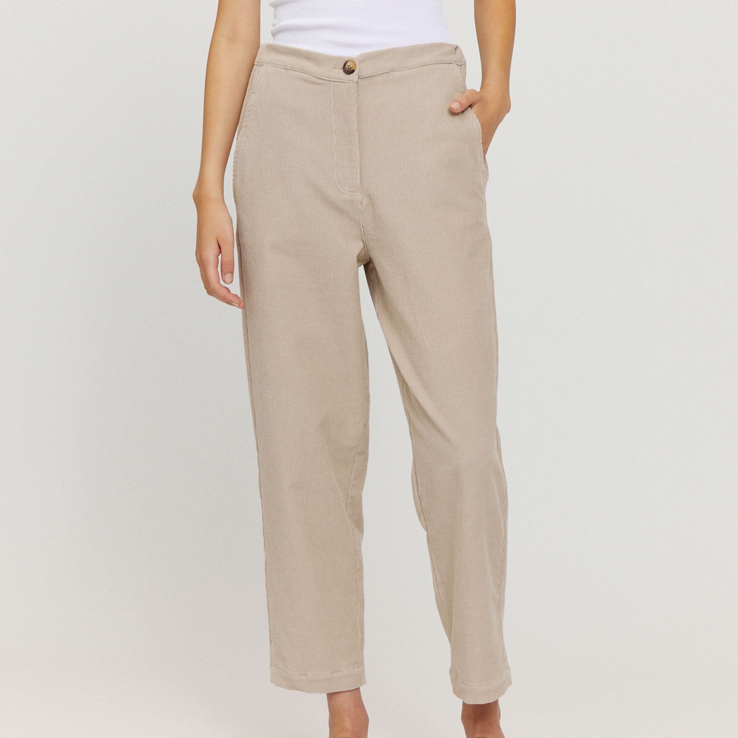 Person in beige high-waisted pants with hand in pocket, wearing a white top, poses against a plain background.